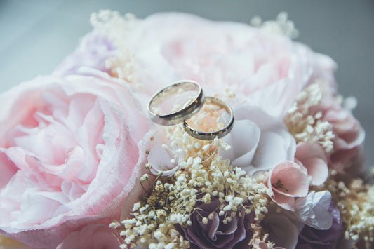 **Alt tag:** Close-up of two wedding rings resting on a pastel floral bouquet with pink roses and delicate white flowers